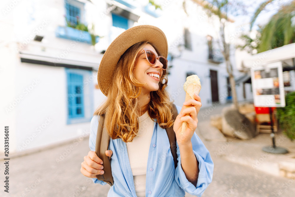 Fototapeta premium A happy tourist in a hat, holding an ice cream cone, strolls along the sunny streets of the city. The young woman admires the city's architecture and eats ice cream. Concept: tourism, vacation.
