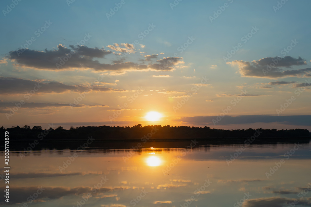 Obraz premium Sunset sky over calm river with golden reflection and dark tree line at horizon, peaceful evening landscape