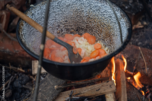 Vegetables simmering in an enamel pot over a campfire in the forest. Cozy outdoor cooking atmosphere featuring fire flames, wooden spoon, rustic style, and traditional camping experience.
