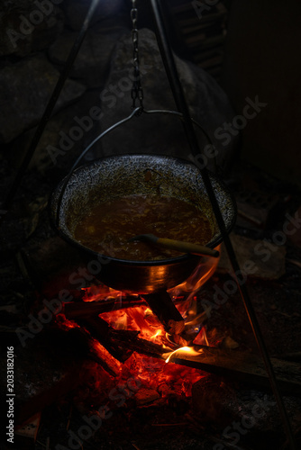 Close-up of carrots and onions being cooked in a hanging pot above an open fire in nature. Authentic camp cooking scene with rustic cookware, flames, smoke, and outdoor survival lifestyle concept.
