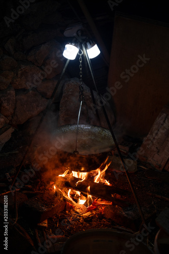 Cooking vegetables in a traditional enamel pot over an open campfire outdoors. Rustic outdoor cooking scene with carrots, onions, firewood, smoke, and camping atmosphere.