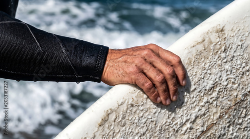 Close-up of a surfer's hand in a black wetsuit gripping a white surfboard with textured wax, with ocean waves in the background.