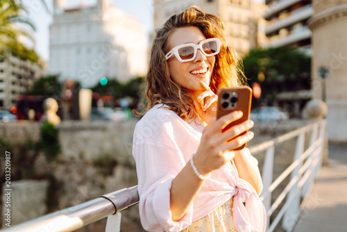 A young woman, holding a phone, strolls through a sunny city near a park. A beautiful woman enjoys the sunset in a stylish dress and with a smartphone. Concept of fashion, style, and relaxation.