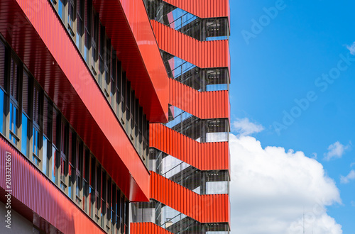 Modern building facade made of red corrugated metal sheet and an outdoor zigzag emergency staircase shot from a low angle, glass windows reflecting light, blue sky with white clouds.