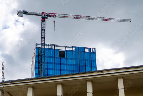 Fotografi A tall white and red construction crane towering over a blue glass skyscraper un