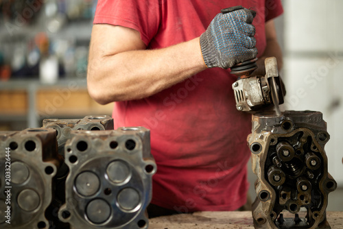 A focused mechanic expertly cleans a cylinder head at a truck maintenance center, utilizing a grinder. The scene captures the precision and skill involved in vehicle maintenance and repair.