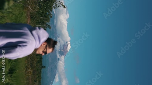Young man in purple hoodie and with bun on her head admires spectacular view of volcano with snow-capped peak and cloud-shrouded slopes. Slow motion.