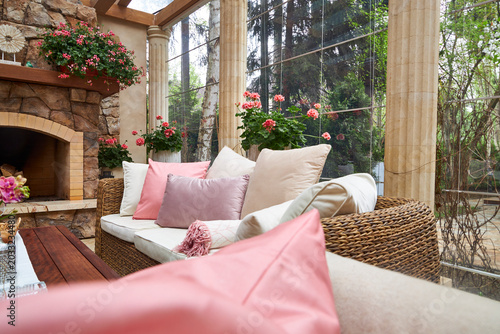 Tableau sur toile Elegant conservatory lounge with wicker furniture, soft pastel cushions and a rustic stone fireplace surrounded by blooming flowers and panoramic forest views