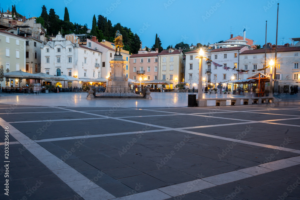 Obraz premium Tartini Square in Piran, Slovenia at Blue Hour with Historic Venetian Architecture and Illuminated Cafes Surrounding the Famous Monument. Evening Scene in the Heart of the Adriatic Coastal Town