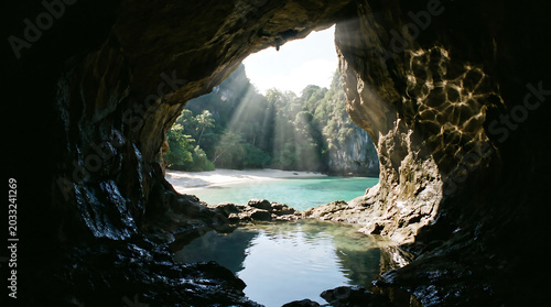 Sunlight pierces through a dark cave opening, illuminating a stunning tropical beach with turquoise waters and lush green cliffs beyond.