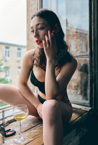 Beautiful young woman sitting on windowsill with red lipstick, old vintage camera and white wine glass. Portrait of gorgeous sad girl in black lingerie