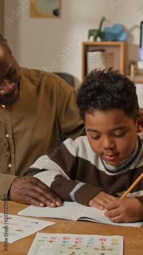 Medium vertical shot of supportive mature African American father encouraging little son during handwriting practice, gently patting on head, while helping with elementary school homework