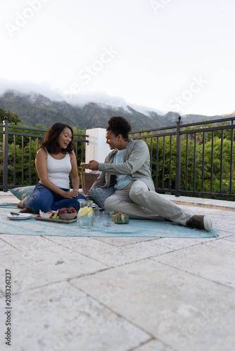 Couple sitting on blue picnic blanket on rooftop terrace exchanging small gift near lemon pitcher