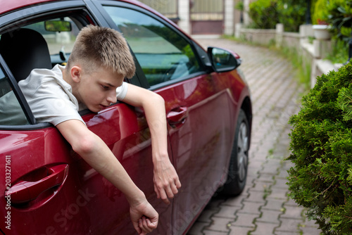 a teenage boy leans out of a red car window looking down with a calm expression and both arms hanging beside the car door outdoors