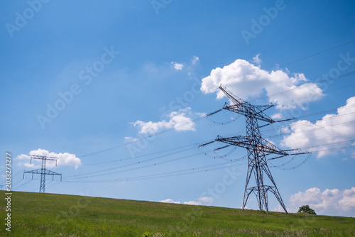 High-voltage electricity pylons and power lines crossing green meadow under blue sky with clouds in rural landscape.
