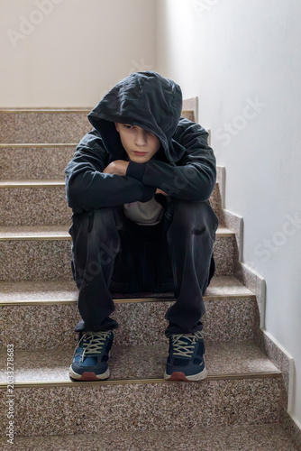 Depressed teenage boy sitting alone on stairs with hood covering face, feeling sad and isolated, youth mental health concept