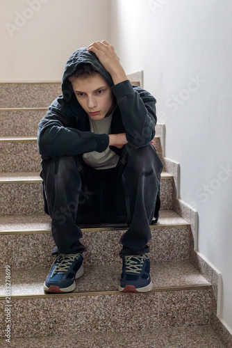 Sad teenage boy sitting on stairs in hoodie, feeling lonely and depressed, youth mental health concept