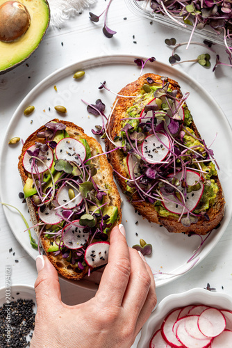 Above view of a hand holding an avocado toast topped with fresh ingredients.
