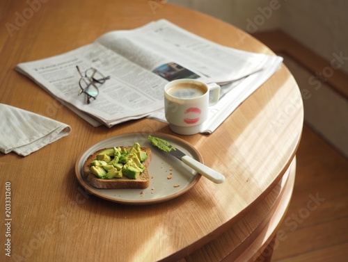 Breakfast setting with avocado toast on plate coffee mug and newspaper on round table.