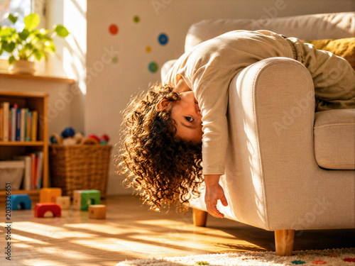 Playful toddler with curly hair hanging upside down from a beige sofa in a warm, sunlit living room with wooden toys on the floor.