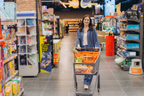 Foto Young woman pushing shopping cart filled with groceries, smiling while walking t
