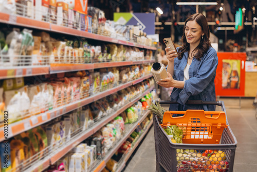 Schilderij op canvas Woman in supermarket aisle checking product information on smartphone while groc