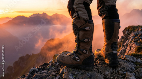 Obraz na plátně Motorcycle adventurer standing on a rocky mountain peak at sunset wearing sturdy
