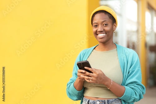 Young woman wearing yellow cap using smartphone and smiling at camera against yellow wall. Woman happy and relaxed. Positive emotion