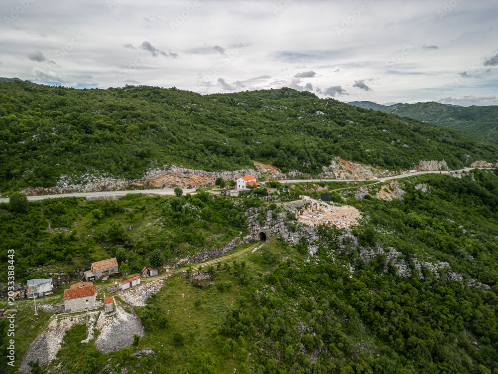 Fototapeta premium Slansko Lake, Montenegro -05 29 2025: Drone landscape photo of hilly terrain in Ponikvica, showing winding road, rural houses with red roofs, rocky outcrops, tunnel entrance, and a quarry under clouds
