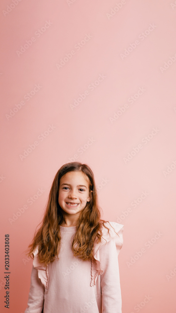 Fototapeta premium Cheerful young girl with long brown hair smiling confidently, standing on a solid pink background, showing joy