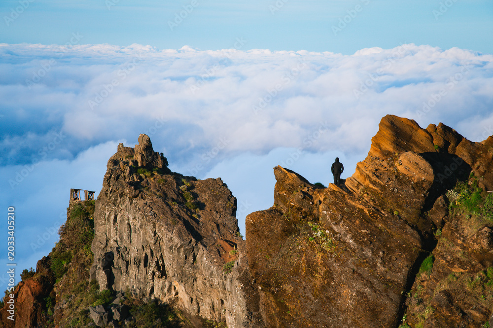 Fototapeta premium A distant silhouette of a hiker standing on a rugged volcanic peak above a vast sea of thick morning clouds.