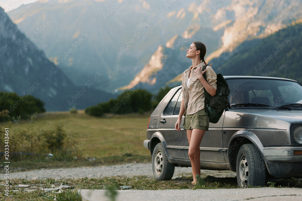 Fototapeta premium Young woman with backpack enjoying a mountain landscape beside a parked car, gazing into the distance. A carefree outdoor lifestyle captures adventure, travel, and nature in warm, soft light on