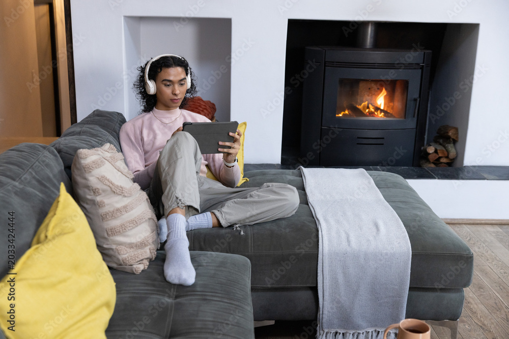 Fototapeta premium Nonbinary person sitting on gray sofa in living room, holding tablet with headphones near fireplace