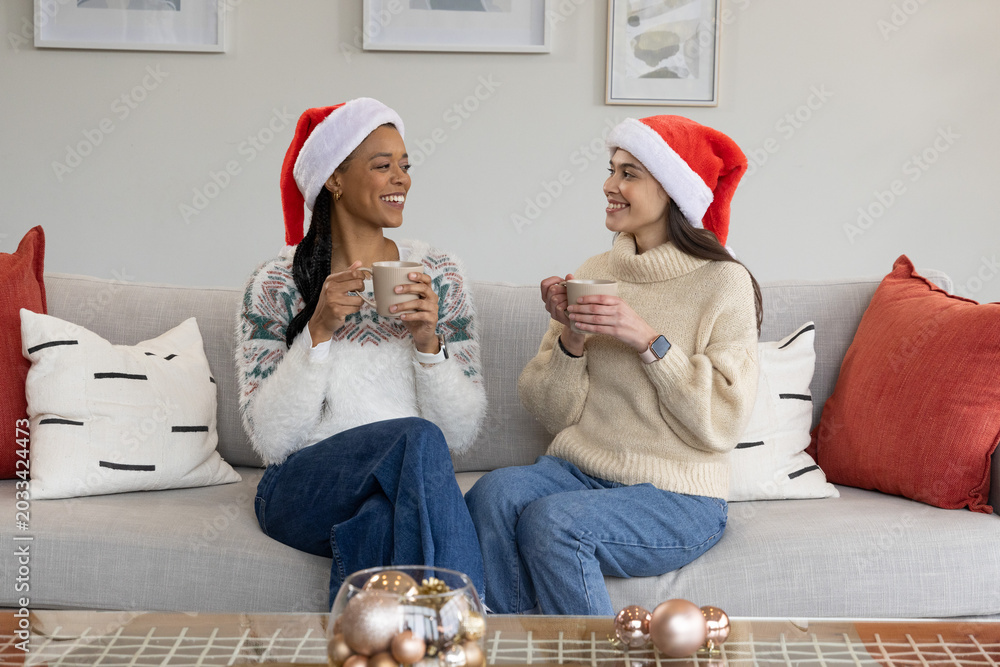 Fototapeta premium African American and Asian women chatting on gray sofa wearing Santa hats, holding ceramic mugs