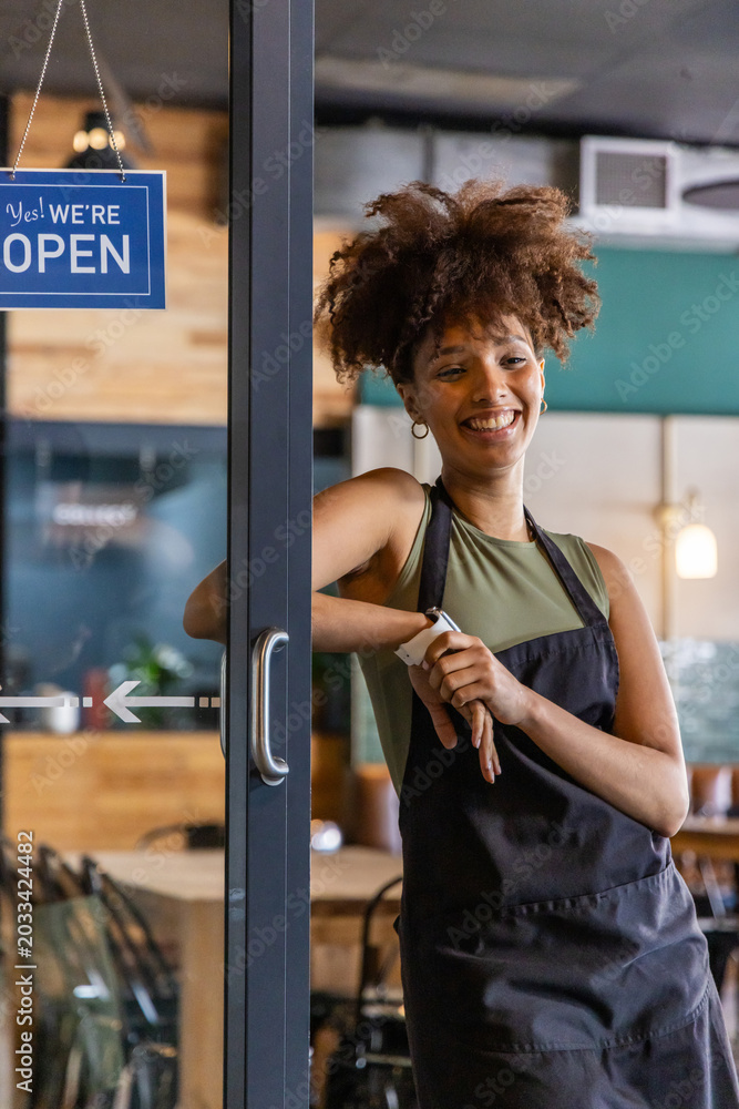Obraz premium African American adult woman smiling and adjusting watch in cafe doorway with black apron OPEN sign