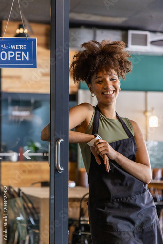 African American adult woman smiling and adjusting watch in cafe doorway with black apron OPEN sign