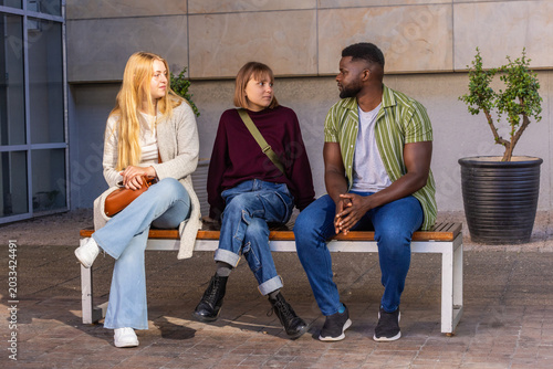 Friends sitting on wooden bench by tiled wall, chatting while holding brown handbag and green bag