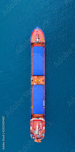 Outstanding vertical overhead drone photograph emphasizing the massive scale of a modern blue and red industrial cargo ship traversing the deep blue ocean surface on a quiet international route