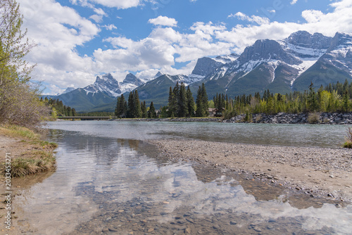 Stunning summer landscape of the Canadian Rocky Mountains and a calm river in Canmore, Alberta. Near Banff National Park with towering mountain peaks, Three Sisters. 