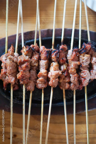 Grilled meat skewers rest on a dark, oval plate. Wooden surface shows natural grain and warm tones. Each skewer holds tender, slightly charred pieces. Fragrant, smoky aroma evokes appetite and comfort