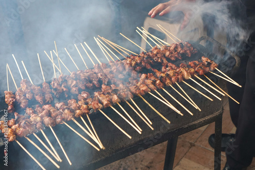 Smoke curls from skewered meat on a grill. Wooden sticks hold chunks of seasoned meat. Grill sits on a dark metal table. Steam rises as food cooks over open flame