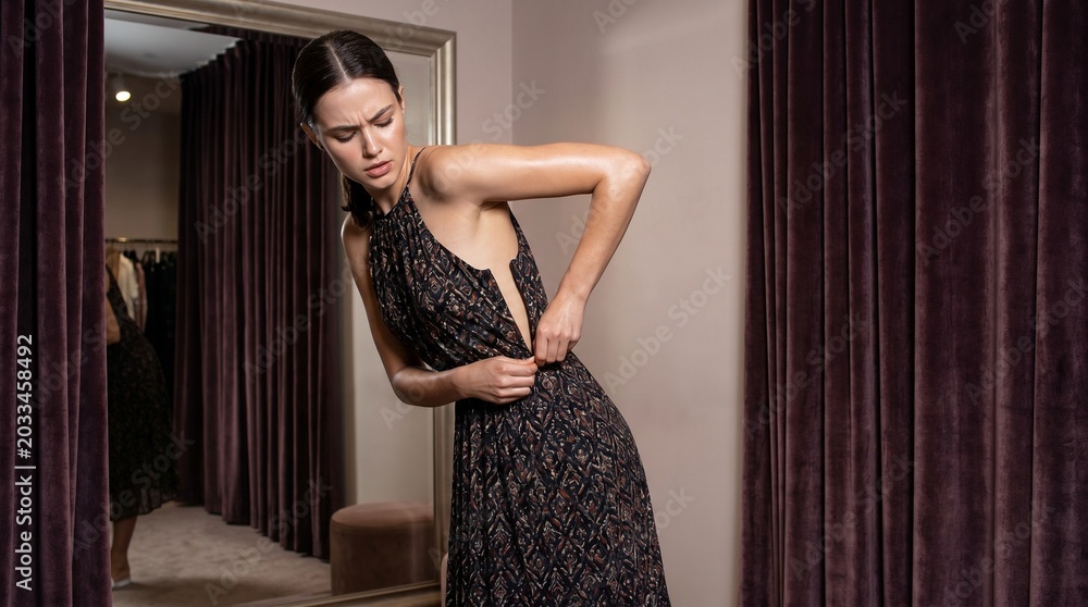 Fototapeta premium Frustrated young woman struggling to zip up tight evening formal dress inside boutique clothing store dressing room