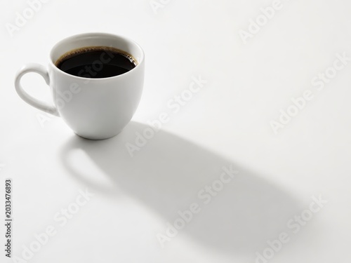 Single white porcelain cup filled with black coffee long diagonal shadow stretching across the white surface clean morning light concept isolated on white seamless background soft directional