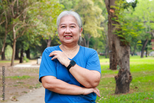Happy senior Asian woman in blue shirt standing confidently in the park. Active elderly lifestyle, health, and retirement concept.