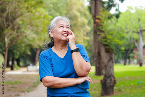 Happy senior Asian woman thinking and smiling in the park. Healthy elderly lifestyle, retirement, and positive mental health concept.