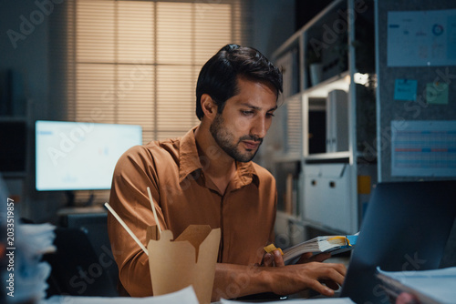 Young Latin businessman eating takeout instant noodles and reviewing document on laptop at desk in dark night office. Overtime working late, overworked deadline, fast food, corporate problem concept.