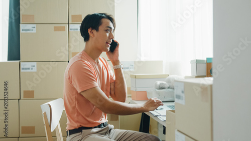 Valokuva Young Asia male entrepreneur small business start up call smartphone confirm order with customer confirm check stock inventory product ready for delivery shipment on laptop at online retail shop