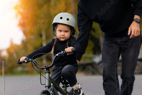 Father teaching young caucasian daughter to ride bicycle outdoors against backdrop of military equipment and tanks. Concept truce and settlement of war, military regime
