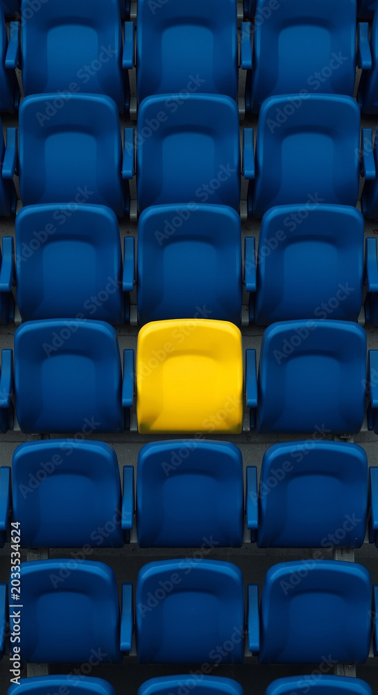 Obraz premium A single yellow stadium seat standing out in a sea of blue seats symbolizing individuality and uniqueness in a crowded public venue.