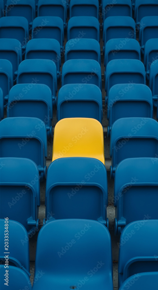 Obraz premium A solitary yellow theater seat stands out among rows of blue seats in a crowded auditorium or stadium symbolizing individuality and uniqueness in a sea of conformity.
